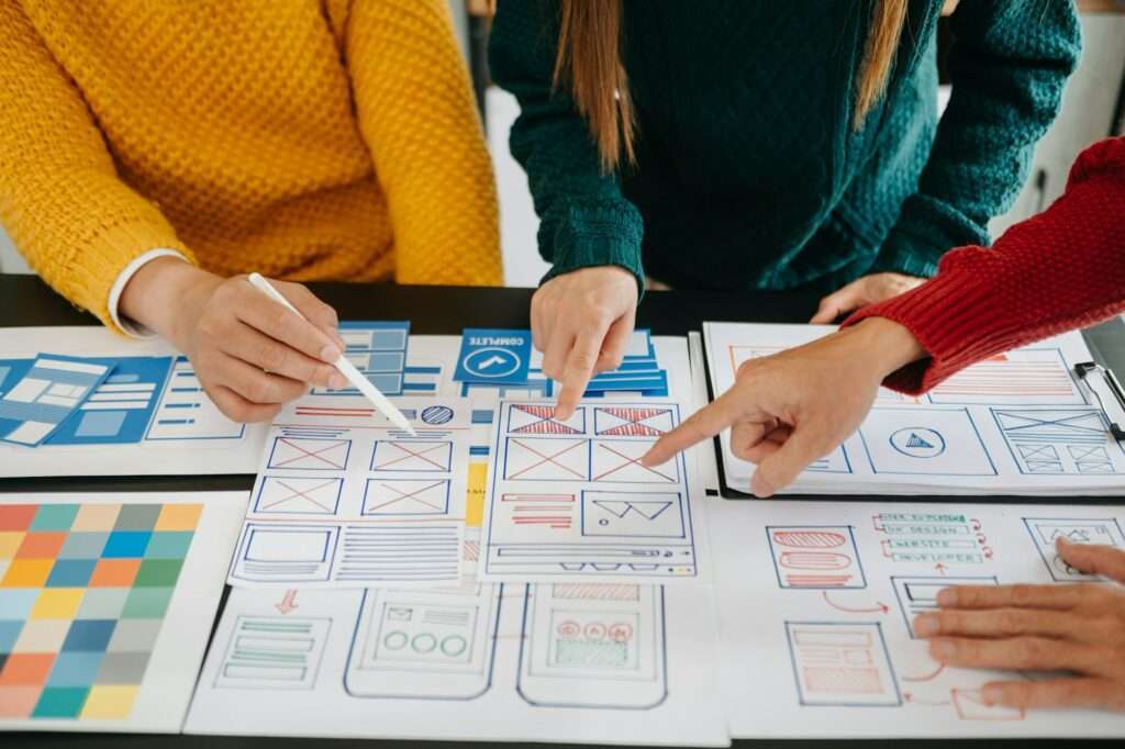 SEO Hannover - Drei Personen arbeiten gemeinsam an einem Tisch und zeigen auf UI/UX-Designskizzen von Website-Layouts. Die Skizzen enthalten detaillierte Wireframes und innovative Ideen. Sie tragen bunte Pullover in Gelb, Rot und Grün. Verschiedene Diagramme und Notizen sind über den Tisch verteilt. Local SEO Agentur Hannover.