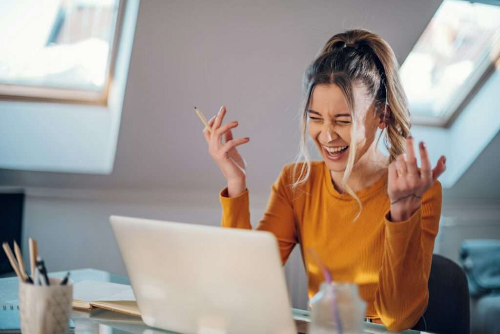 SEO Hannover - Eine Person mit zusammengebundenen langen Haaren und einem gelben Pullover sitzt an einem Schreibtisch und ist in die Erstellung einer Webvisitenkarte vertieft. Sie lächelt und gestikuliert mit einem Stift in der Hand. Natürliches Licht fällt durch schräge Fenster und beleuchtet die auf dem Schreibtisch verstreuten Kunstutensilien. Local SEO Agentur Hannover.