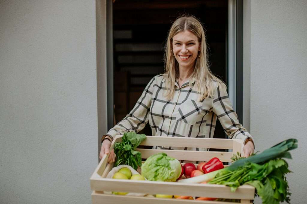 SEO Hannover - Eine Person steht in einer Tür und hält eine Holzkiste mit frischem Gemüse, darunter Salat, Paprika, Lauch und Äpfel. In ihrem karierten Hemd lächelt sie und ähnelt einem Bild aus einem Stockfoto von Google SEO. Local SEO Agentur Hannover.