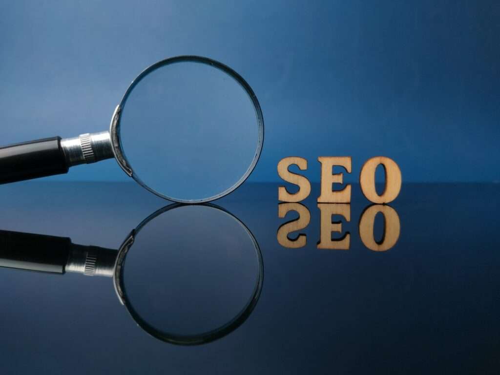 SEO Hannover - Neben den Holzbuchstaben „SEO“ auf einer reflektierenden blauen Oberfläche befindet sich eine Lupe, wodurch ein Spiegeleffekt entsteht, der an die für Google SEO erforderliche Präzision erinnert. Local SEO Agentur Hannover.