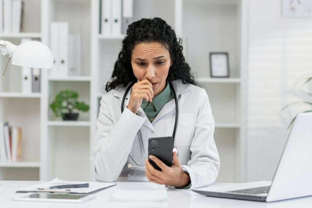 SEO Hannover - Eine Ärztin mit langen Locken sitzt besorgt an einem Schreibtisch, während sie auf ihrem Smartphone Online-Bewertungen überprüft. Papiere, ein Stift, ein Laptop und eine Schreibtischlampe sind zu sehen. Sie trägt einen weißen Kittel und ein Stethoskop und fragt sich, wie sie Google-Bewertungen effektiv verwalten kann. Local SEO Agentur Hannover.