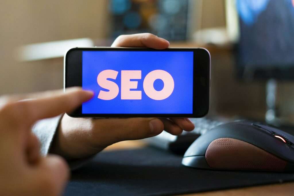 SEO Hannover - Eine Person hält ein Smartphone, auf dem in großen weißen Buchstaben auf blauem Hintergrund das Wort „SEO“ steht. Das Telefon, das die Expertise einer SEO Agentur Hannover symbolisiert, liegt über einer Computermaus auf einem Schreibtisch, mit unscharfem Hintergrund. Local SEO Agentur Hannover.