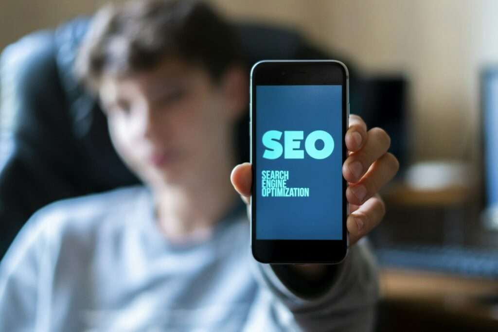 SEO Hannover - Eine Person hält ein Smartphone, auf dessen Bildschirm der Text „SEO Suchmaschinenoptimierung“ angezeigt wird. Der Hintergrund ist unscharf und der Fokus liegt auf dem Telefon. Local SEO Agentur Hannover.