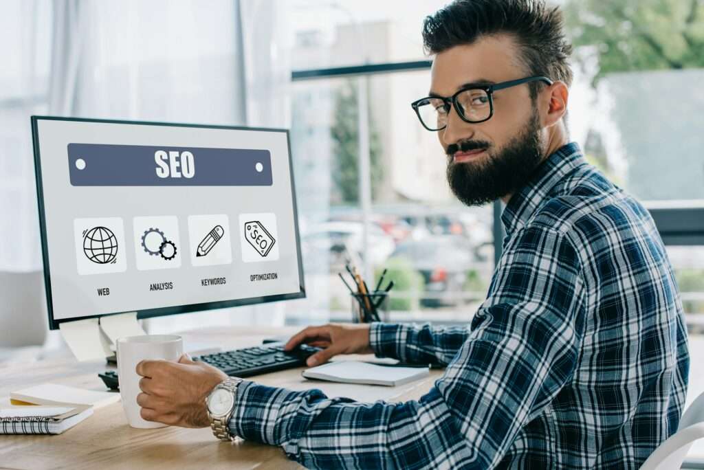 SEO Hannover - Eine Person sitzt an einem Schreibtisch in einem Büro und konzentriert sich auf einen Computerbildschirm, auf dem eine Google SEO-Oberfläche mit Symbolen wie „Web“, „Analyse“, „Keywords“ und „Optimierung“ angezeigt wird. Die Person hält eine Tasse, trägt eine Brille und ein kariertes Hemd. Local SEO Agentur Hannover.