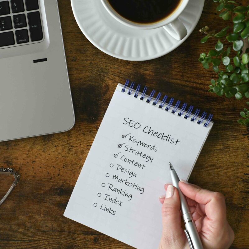 Webdesign Hannover - Eine Hand hält einen Stift und schreibt auf einen Notizblock mit der Überschrift „SEO-Checkliste“ und listet Punkte wie Schlüsselwörter, Strategie, Inhalt, Design, Marketing, Ranking, Index und Links auf. In der Nähe stehen ein Laptop, eine Tasse Kaffee, Gläser und eine Pflanze auf dem Holztisch. Local SEO Agentur Hannover.