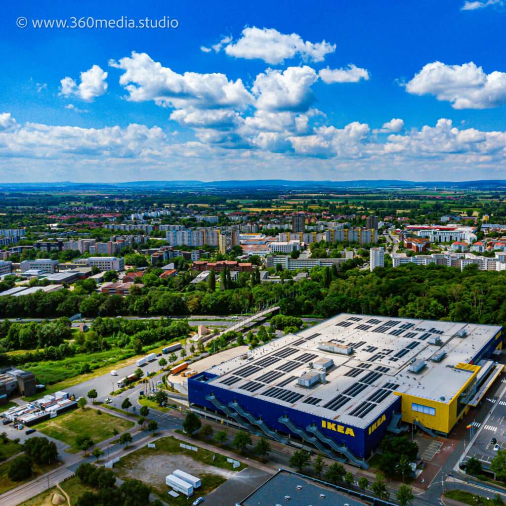 Webdesign Hannover - Drohnenaufnahmen aus der Luft zeigen ein mit Solarmodulen ausgestattetes IKEA-Geschäft inmitten eines belebten Parkplatzes. Im Hintergrund entfaltet sich eine lebendige Stadtlandschaft mit Wohnhäusern, üppigen Grünflächen und einem blauen Himmel mit vereinzelten Wolken. Local SEO Agentur Hannover.