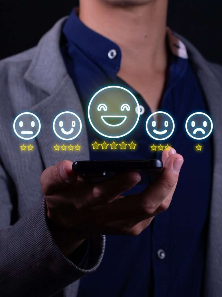 Webdesign Hannover - Eine Person im Anzug hält ein Smartphone, auf dem eine Reihe schwebender digitaler Symbole mit Smileys und Sternebewertungen von traurig bis glücklich angezeigt werden. Das Highlight ist das glückliche Gesicht mit fünf Sternen, das symbolisiert, wie Unternehmen ihr Image verbessern können, indem sie Google-Bewertungen auf ihre Website einbinden. Local SEO Agentur Hannover.