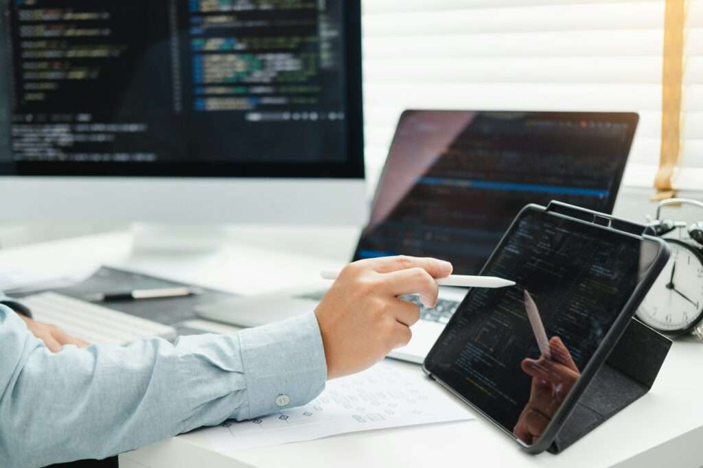 Webdesign Hannover - Eine Person benutzt einen Stift auf einem Tablet, auf dem Code angezeigt wird, und denkt über die Frage nach: „Ist WebP besser als JPG?“ In der Nähe zeigen ein Desktop-Monitor und ein Laptop ebenfalls Programmierung. Papiere und eine Uhr zieren den Schreibtisch und schaffen eine Umgebung, in der es um Codierung und Softwareentwicklung geht. Local SEO Agentur Hannover.