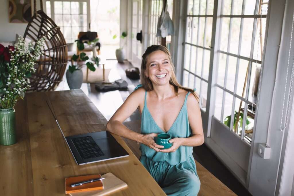 Webdesign Hannover - Eine Frau in einem hellblauen Kleid sitzt an einem Holztisch, hält eine Tasse und lächelt. Ein Laptop und ein Notizblock mit einem Stift liegen daneben, während sie überlegt: „Wie kann ich Keywords finden?“ Der Raum ist durch große Fenster in natürliches Licht getaucht und mit Topfpflanzen geschmückt. Im Hintergrund steht ein runder Holzstuhl. Local SEO Agentur Hannover.