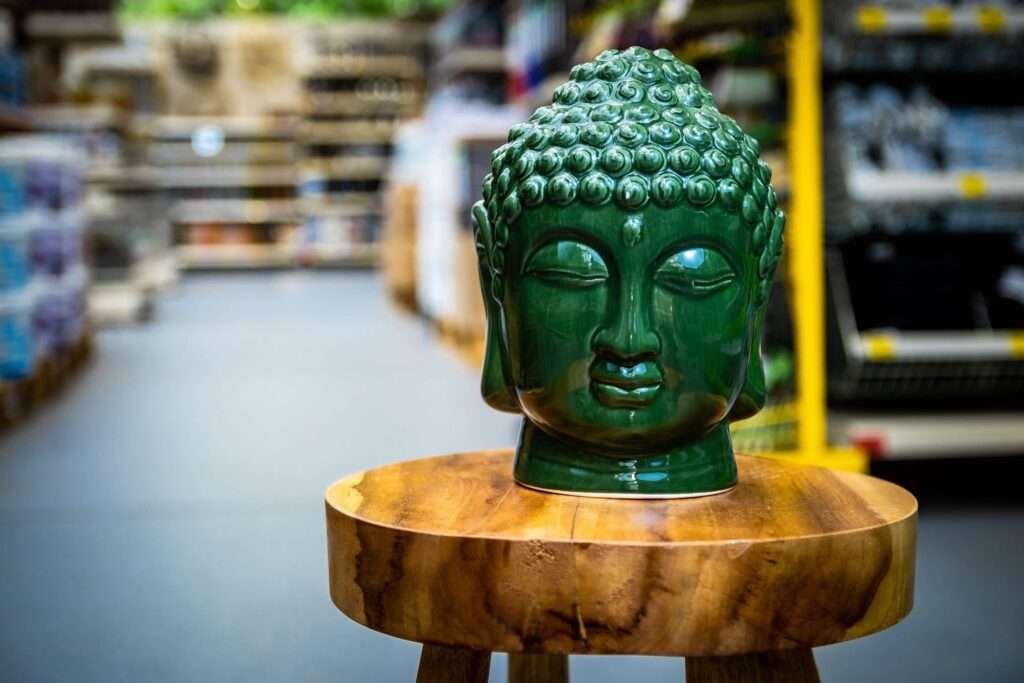 Webdesign Hannover - Ein grüner Buddhakopf aus Keramik sitzt friedlich auf einem Holzhocker in einem Ladengang, wunderschön eingefangen in dieser Fotografie. Regale mit verschiedenen Gegenständen verschwimmen sanft im Hintergrund und verleihen der Szene Tiefe. Local SEO Agentur Hannover.
