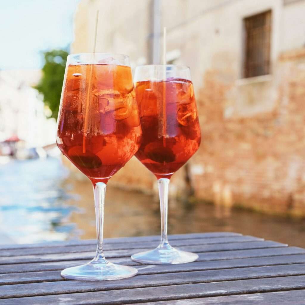 SEO Hannover - Zwei hohe Gläser Aperol Spritz, die über Eiswürfeln klirren, mit leuchtenden Orangenscheiben garniert und auf einem Holztisch platziert sind. Im unscharfen Hintergrund runden ein malerischer Kanal und charmante Backsteingebäude diese Szene ab, die sich perfekt zur Verbesserung von SEO für Bilder eignet. Local SEO Agentur Hannover.