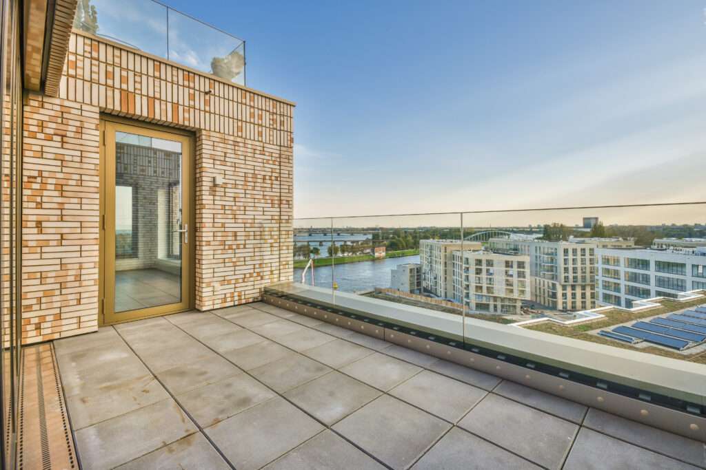 SEO Hannover - Eine Dachterrasse, die sich perfekt für jeden Immobilienfotografen eignet, verfügt über ein Glasgeländer mit Blick auf moderne Gebäude und einen Fluss. Der großflächig geflieste Boden ergänzt die Szene, während eine Tür mit Glasfenster in das charmante Gebäude mit Backsteinmauern führt. Der Himmel ist klar und hell. Local SEO Agentur Hannover.