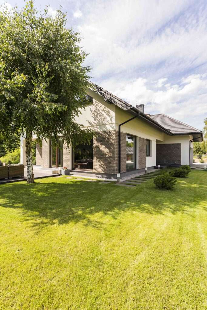 SEO Hannover - Ein modernes einstöckiges Haus mit hellen Wänden und einem dunklen Ziegeldach ist von einem üppigen, grünen Rasen umgeben. Links steht ein Baum, der das Haus teilweise beschattet. Perfekt für Immobilienfotografie, der Himmel ist klar mit ein paar Wolken. Local SEO Agentur Hannover.