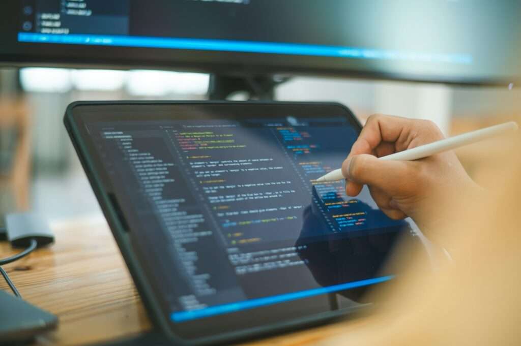 SEO Hannover - Eine Person verwendet einen Stift auf einem Tablet-Bildschirm, auf dem Code angezeigt wird, während ein großer Monitor mit weiteren Codezeilen den Hintergrund bildet. Der Arbeitsbereich suggeriert eine Umgebung, in der der Schwerpunkt auf Programmierung oder der Erforschung komplexer Themen wie dem Unterschied zwischen Template und Theme in der Softwareentwicklung liegt. Local SEO Agentur Hannover.