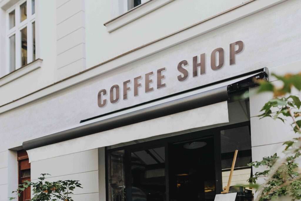 Webdesign Hannover - Ein weißes Gebäude rühmt sich eines Schilds mit der eleganten Aufschrift „Coffee Shop“, das ein durchdachtes Logodesign widerspiegelt. Über dem Eingang ergänzt eine schicke, einziehbare schwarz-weiße Markise das Ambiente, während Grünpflanzen im Vordergrund einen Hauch von Natur verleihen. Local SEO Agentur Hannover.