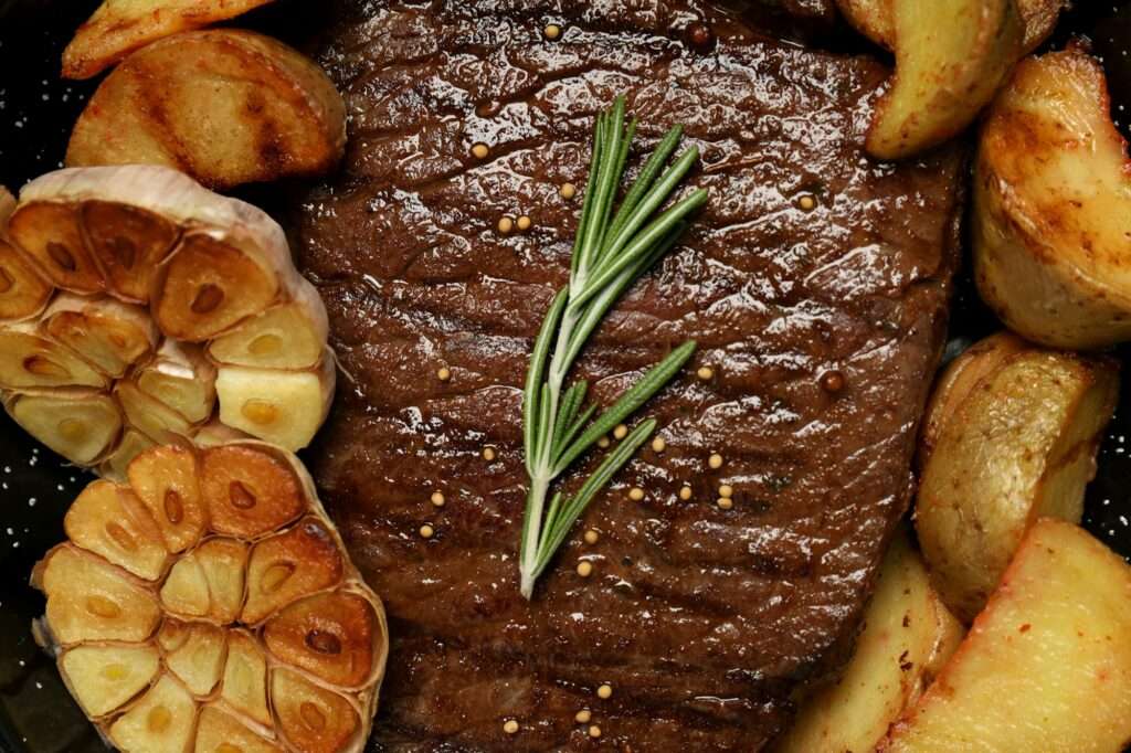 SEO Hannover - Eine Nahaufnahme eines gebratenen Steaks, garniert mit einem Zweig Rosmarin, perfekt gestylt für SEO für Bilder. Das Steak ist von gerösteten Kartoffelspalten und einer halbierten Knolle gerösteten Knoblauchs umgeben. Local SEO Agentur Hannover.