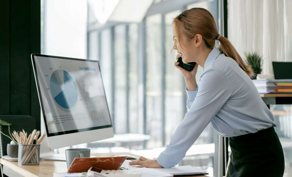 Confident businesswoman using mobilphone and computer analyzing business growth graph online.