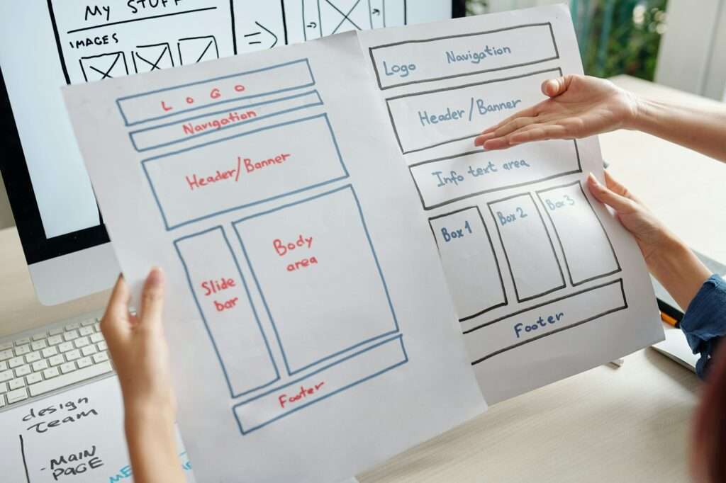 SEO Hannover - Zwei Hände halten Skizzen des Website-Layouts auf Papier, die Abschnitte wie Logo, Navigationsmenü, Kopfzeile, Banner und Fußzeile zeigen. Ein Computerbildschirm im Hintergrund spiegelt das Website-Design wider. Verschiedene Notizen und Diagramme sind über den Arbeitsbereich verstreut. Local SEO Agentur Hannover.