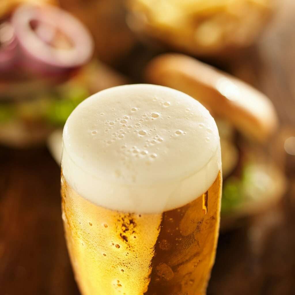 SEO Hannover - Eine Nahaufnahme eines gekühlten, goldenen Biers in einem Glas mit Schaumkrone. Im unscharfen Hintergrund sind Andeutungen eines Sandwichs mit Zwiebelringen und einer Schale Pommes zu erkennen, die die Essenz eines zwanglosen Essens einfangen. Dieses Bild verkörpert sowohl Geschmack als auch Ambiente, perfekt für SEO für Bilder-Enthusiasten, die nach visueller Attraktivität suchen. Local SEO Agentur Hannover.