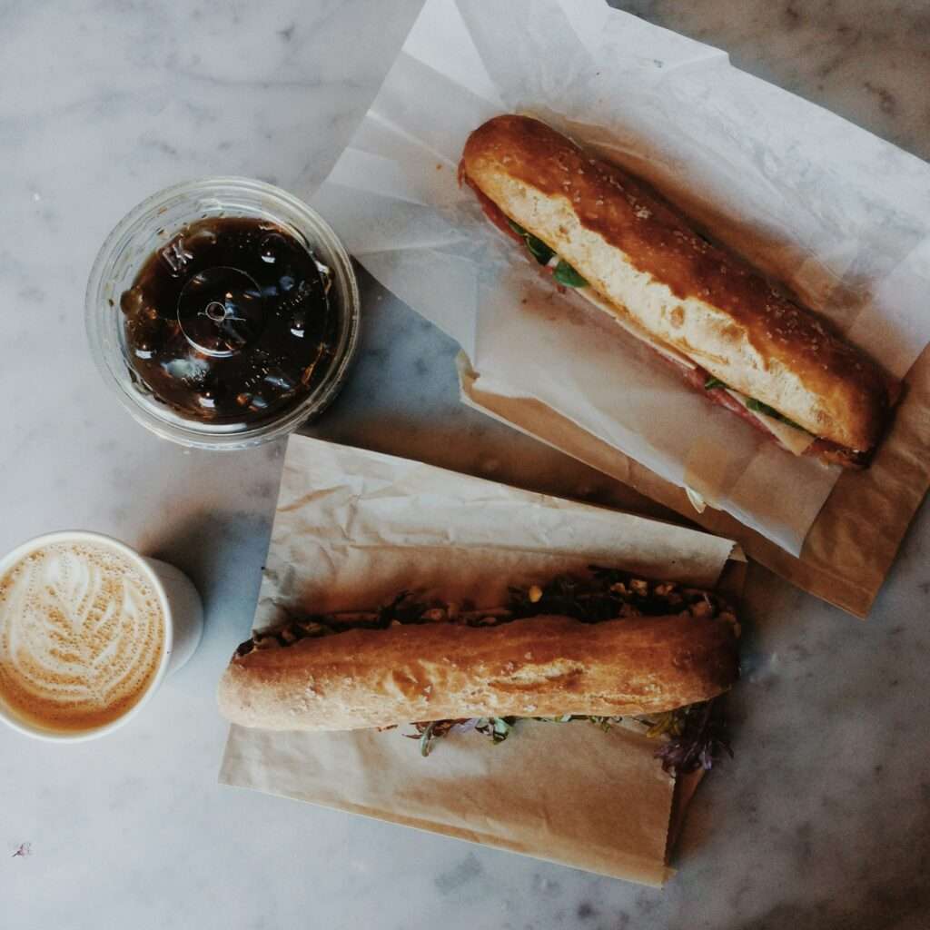 SEO Hannover - Zwei Sandwiches liegen auf braunem Papier auf einer Marmoroberfläche. Links steht eine durchsichtige Tasse mit Eiskaffee, während ein Latte mit Schaumkunst die untere linke Ecke ziert – eine perfekte Anordnung für die Gestaltung optisch ansprechender Bilder, die die SEO für Bilder verbessern. Local SEO Agentur Hannover.