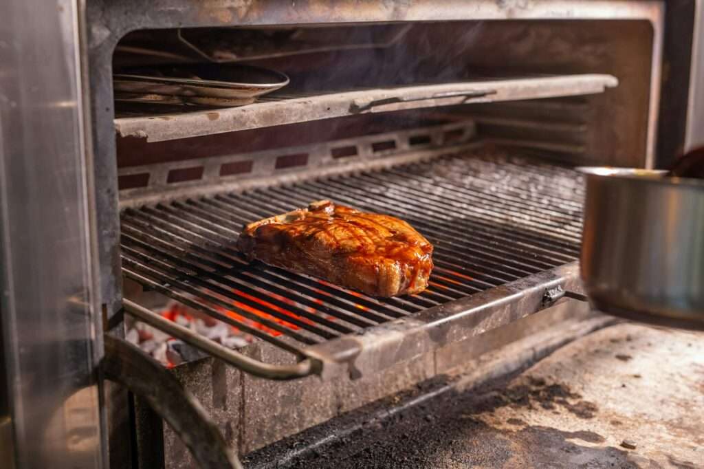 SEO Hannover - Ein dickes Steak wird in einem Ofen gegrillt, brutzelnd und rauchend, als ob SEO für Bilder jedes Detail optimiert hätte. Die Grillstäbe sind erhitzt und das Steak scheint über glühender Glut zu garen, mit sichtbaren Grillspuren und einer glänzenden Beschichtung. Auf der rechten Seite ist teilweise ein Topf sichtbar. Local SEO Agentur Hannover.