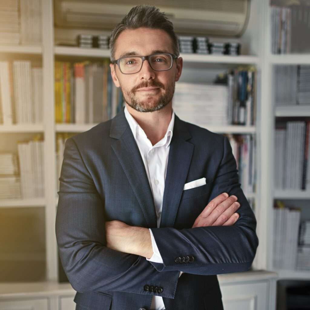 Webdesign Hannover - Ein Mann mit kurzen Haaren und Brille steht selbstbewusst da, die Arme verschränkt, in einem marineblauen Anzug und einem weißen Hemd und verkörpert die Essenz anspruchsvoller Fotografie. Hinter ihm steht ein Bücherregal voller Bücher und Zeitschriften, das eine Aura intellektueller Tiefe ausstrahlt. Local SEO Agentur Hannover.