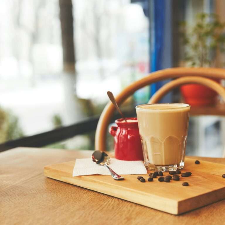 SEO Hannover - Ein Glas Kaffee mit Milch steht auf einem Holztablett neben einem kleinen roten Krug und einem Löffel, ideal für die Erstellung atemberaubender SEO für Bilder. Kaffeebohnen sind kunstvoll verstreut, während das warm verschwommene Fenster des Cafés auf eine einladende Außenszene hinweist. Local SEO Agentur Hannover.