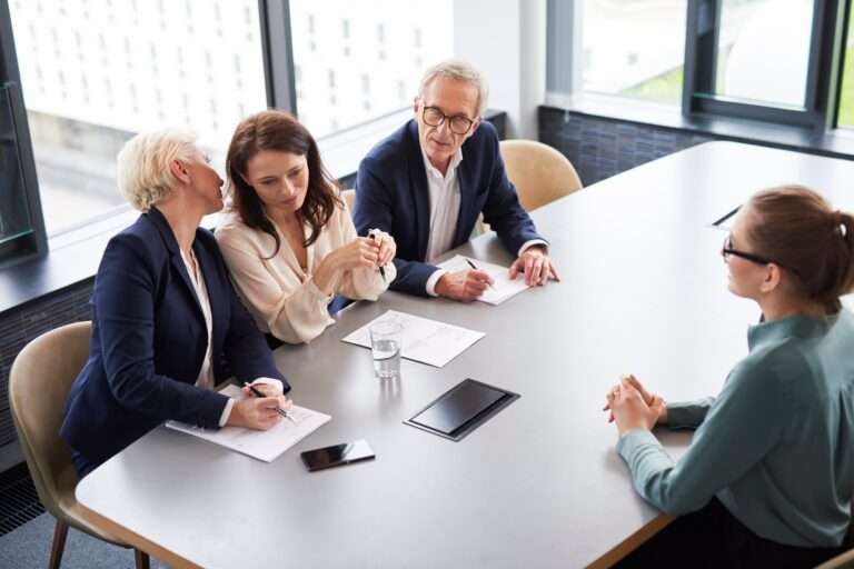 Webdesign Hannover - Eine Person sitzt drei anderen am Konferenztisch gegenüber und bespricht möglicherweise neue Möglichkeiten bei Google4Jobs. Einer flüstert dem anderen etwas zu, während Papiere und Stifte herumliegen. Ein Smartphone und ein Glas Wasser stehen in der Nähe, während große Fenster einen atemberaubenden Blick auf die Stadt bieten. Local SEO Agentur Hannover.