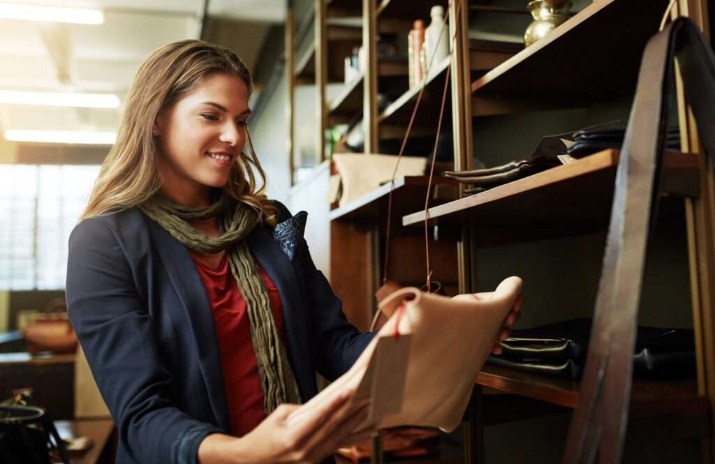 Webdesign Hannover - Eine Frau mit langen Haaren, Schal und Jacke begutachtet neugierig eine beige Tasche im Laden. Sie steht neben einem Regal, auf dem verschiedene Artikel ausgestellt sind, darunter weitere Taschen und Flaschen. Der Laden mit seinen Holzregalen und dem hellen Interieur lädt die Käufer ein, weiter zu stöbern. Local SEO Agentur Hannover.