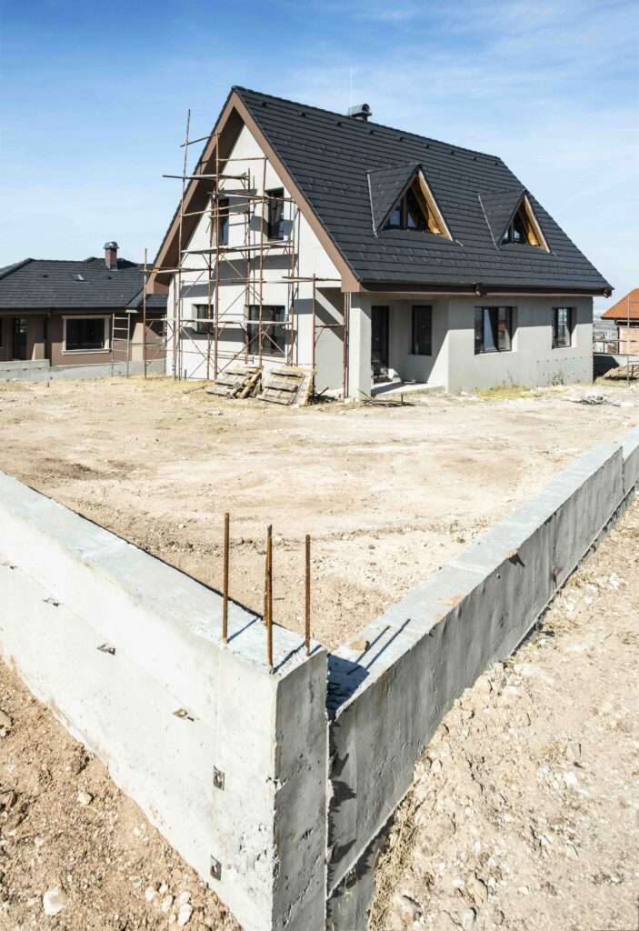 SEO Hannover - Ein teilweise fertiggestelltes Haus mit dunklem Dach und Gerüst auf einer Seite steht auf einem unbefestigten Grundstück, ein hervorragendes Motiv für Immobilienfotografie. In der Nähe ist im Vordergrund unter dem klaren blauen Himmel ein Betonfundament zu sehen. Local SEO Agentur Hannover.