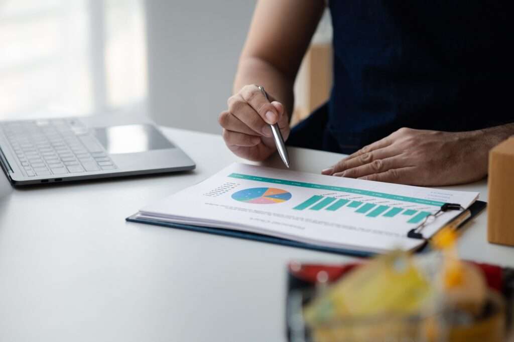 Webdesign Hannover - Eine Person sitzt mit einem Laptop an einem Schreibtisch und denkt über die Frage nach: „Wie viel kostet die Pflege einer Website?“, während sie einen Stift in der Hand hält und einen Bericht mit bunten Grafiken und Diagrammen liest, der an einem Klemmbrett befestigt ist. Im Vordergrund ist ein Korb mit Schreibwaren zu sehen. Local SEO Agentur Hannover.