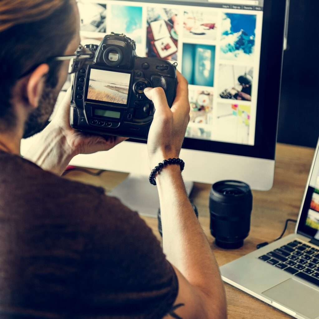 Webdesign Hannover - Eine Person mit Bart und Brille betrachtet Fotos auf einer Digitalkamera und vertieft sich in die Kunst der Fotografie. Sie sitzt vor einem Computermonitor, auf dem verschiedene Bilder angezeigt werden. In der Nähe liegt ein Kameraobjektiv neben einem offenen Laptop auf dem Holzschreibtisch. Local SEO Agentur Hannover.