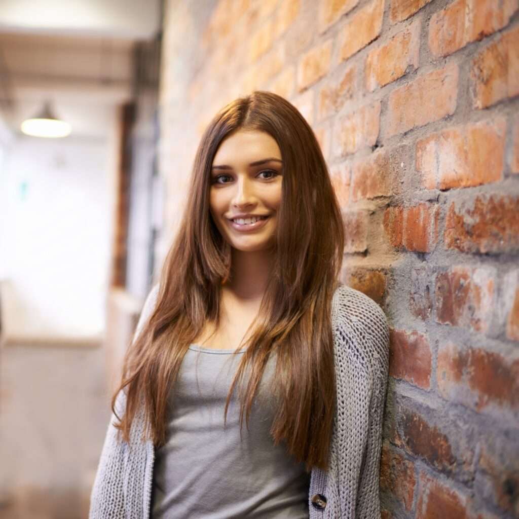 Webdesign Hannover - Eine Person mit langen braunen Haaren steht in einem grauen Pullover und Hemd vor einer Backsteinmauer, festgehalten in einer atemberaubenden Fotografie. Der sanft beleuchtete Innenraum mit einer weiteren Backsteinmauer im Flur verleiht der Szene Tiefe. Local SEO Agentur Hannover.