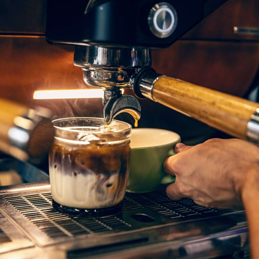 SEO Hannover - Eine Kaffeemaschine gießt Espresso in ein Glas mit Milch und zaubert so ein wunderschön geschichtetes Getränk. Eine Hand hält eine grüne Tasse in der Nähe, bereit für das perfekte Foto, um SEO für Bilder zu verbessern. Der Hintergrund zeigt das warme Leuchten einer braunen Kaffeemaschine. Local SEO Agentur Hannover.