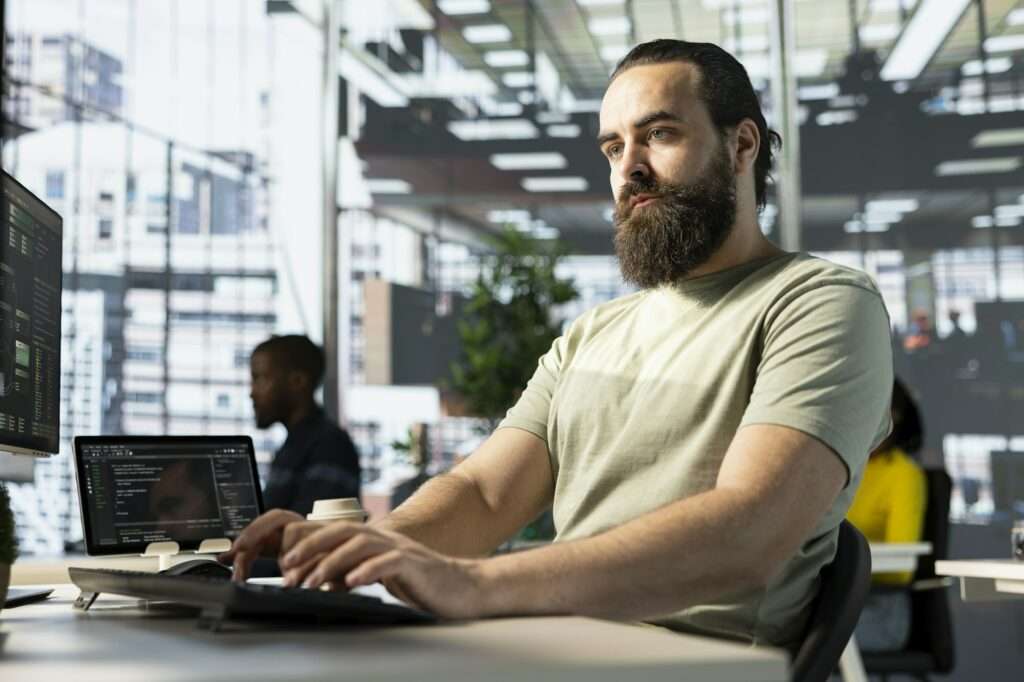 Webdesign Hannover - Ein bärtiger Mann tippt konzentriert Code auf einem Computer in einem modernen Büro. Ein Laptop und zwei Monitore zeigen brillante Codezeilen. Kollegen sind im Hintergrund verschwommen und der Raum erstrahlt in natürlichem Licht, was die Essenz von „Was ist ein gutes Keyword?“ einfängt, während Konzentration die Oberhand gewinnt. Local SEO Agentur Hannover.