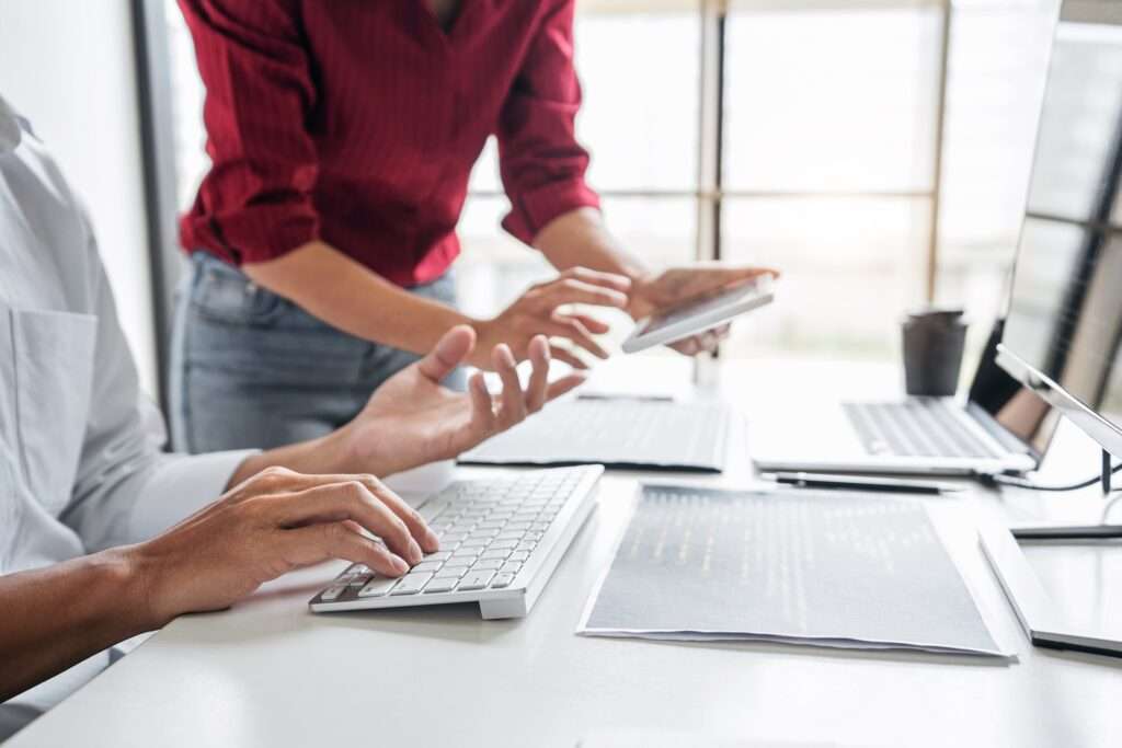 Webdesign Hannover - Zwei Personen arbeiten an einem Schreibtisch gemeinsam an einem Projekt. Einer tippt auf einer Tastatur, während der andere auf ein Tablet zeigt und das Wesentliche dessen bespricht, was auf einer Internetseite vorhanden sein muss. Mehrere Dokumente und Laptops liegen verstreut herum, im Hintergrund ein großes Fenster. Local SEO Agentur Hannover.