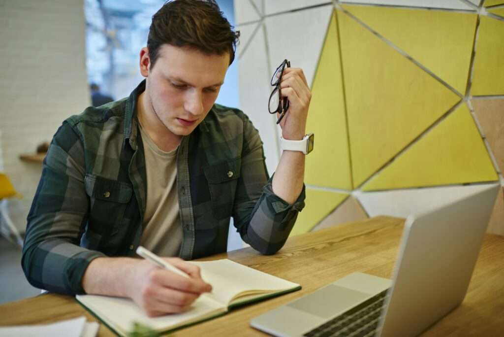 SEO Hannover - Eine Person sitzt an einem Holztisch, hält eine Brille in der Hand und macht sich Notizen für eine Webseite für Steuerberater. Ein geöffneter Laptop fesselt ihre Aufmerksamkeit, während im Hintergrund eine geometrisch gemusterte Wand in Gelb- und Weißtönen zu sehen ist. Die Person wirkt konzentriert und aufmerksam. Local SEO Agentur Hannover.