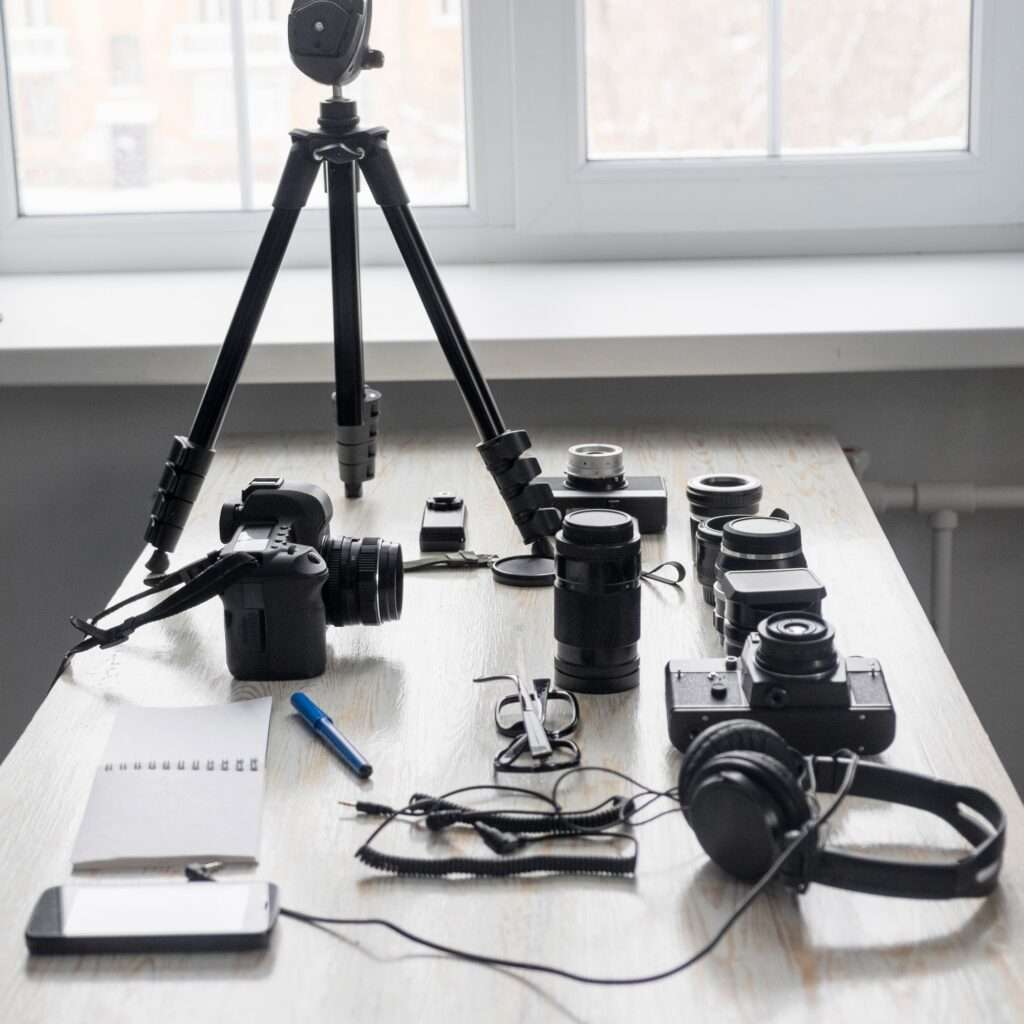 Webdesign Hannover - Ein Tisch voller Fotoausrüstung, darunter ein Stativ, mehrere Kameras, Objektive, ein Notebook und Kopfhörer. Dieser intime Arbeitsbereich von Fotografie wird sanft durch das sanfte Tageslicht beleuchtet, das durch das nahe gelegene Fenster fällt. Local SEO Agentur Hannover.