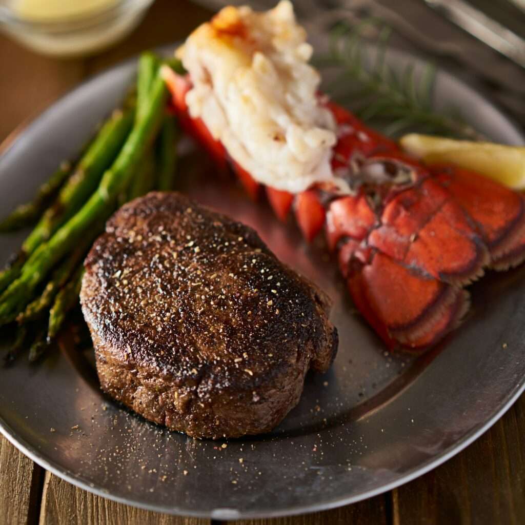 SEO Hannover - Ein mit Pfeffer gewürztes Steak wird elegant auf einem Metallteller zusammen mit einem Hummerschwanz und gegrilltem Spargel serviert. Eine Zitronenscheibe und ein Rosmarinzweig runden dieses optisch beeindruckende Gericht ab, das sich perfekt für SEO-Bilder eignet, die die Sinne verführen. Local SEO Agentur Hannover.