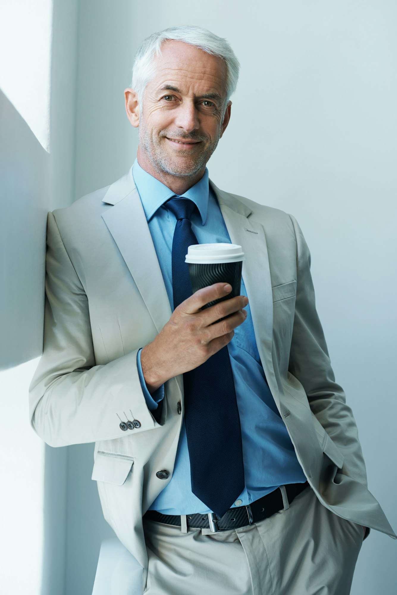 SEO Hannover - Ein vornehmer älterer Mann mit kurzen grauen Haaren, hellblauem Hemd, beigefarbenem Jackett und marineblauer Krawatte lehnt mit einer Kaffeetasse in der Hand an einer weißen Wand. Aufgenommen von Business Fotograf Hannover, strahlt er einen entspannten Gesichtsausdruck aus, eine Hand lässig in der Tasche. Local SEO Agentur Hannover.