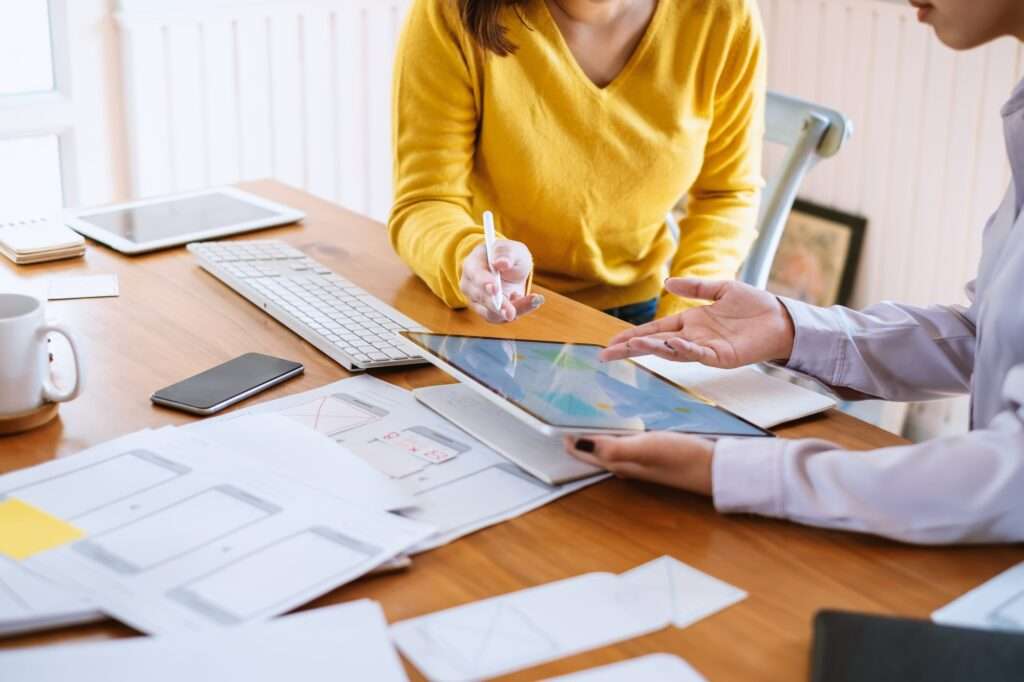 Webdesign Hannover - Zwei Personen an einem Schreibtisch arbeiten mit einem Tablet und einem Stift an einem digitalen Design. Zwischen Papieren mit Skizzen, einer Tastatur und einem Telefon zeigt eine Person im gelben Pullover auf das Tablet. Während sie darüber diskutieren, was „auf einer Internetseite vorhanden sein muss“, gestikuliert die andere nachdenklich. Local SEO Agentur Hannover.