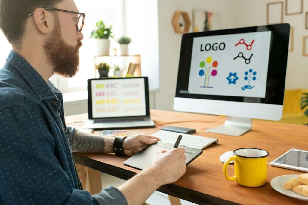 SEO Hannover - Eine bärtige Person ist in die Gestaltung eines Logos vertieft und bedient gekonnt ein Grafiktablett, auf dessen Computerbildschirm Designs leuchten. Auf dem Holzschreibtisch stehen ein Laptop und eine gelbe Tasse. Der Raum wird durch natürliches Licht erhellt, das die Wanddekoration im Hintergrund hervorhebt. Local SEO Agentur Hannover.