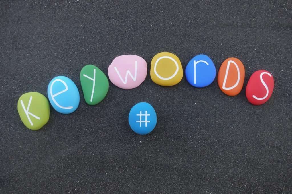 Webdesign Hannover - Auf dunklem Sand sind bunte Steine angeordnet, die das Wort „Keywords“ bilden. In der Mitte befindet sich zusätzlich ein blauer Stein mit einem Hashtag-Symbol, das an die Optimierung von Google Keywords erinnert. Local SEO Agentur Hannover.