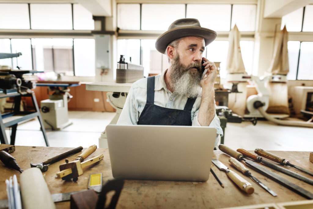 Webdesign Hannover - Ein Mann mit Hut und Schürze sitzt an einer Werkbank in einer Werkstatt und bespricht Handwerkeraufträge auf seinem Smartphone. Vor ihm liegt ein geöffneter Laptop inmitten verstreuter Holzbearbeitungswerkzeuge. Große Fenster im Hintergrund durchfluten den Raum mit natürlichem Licht. Local SEO Agentur Hannover.