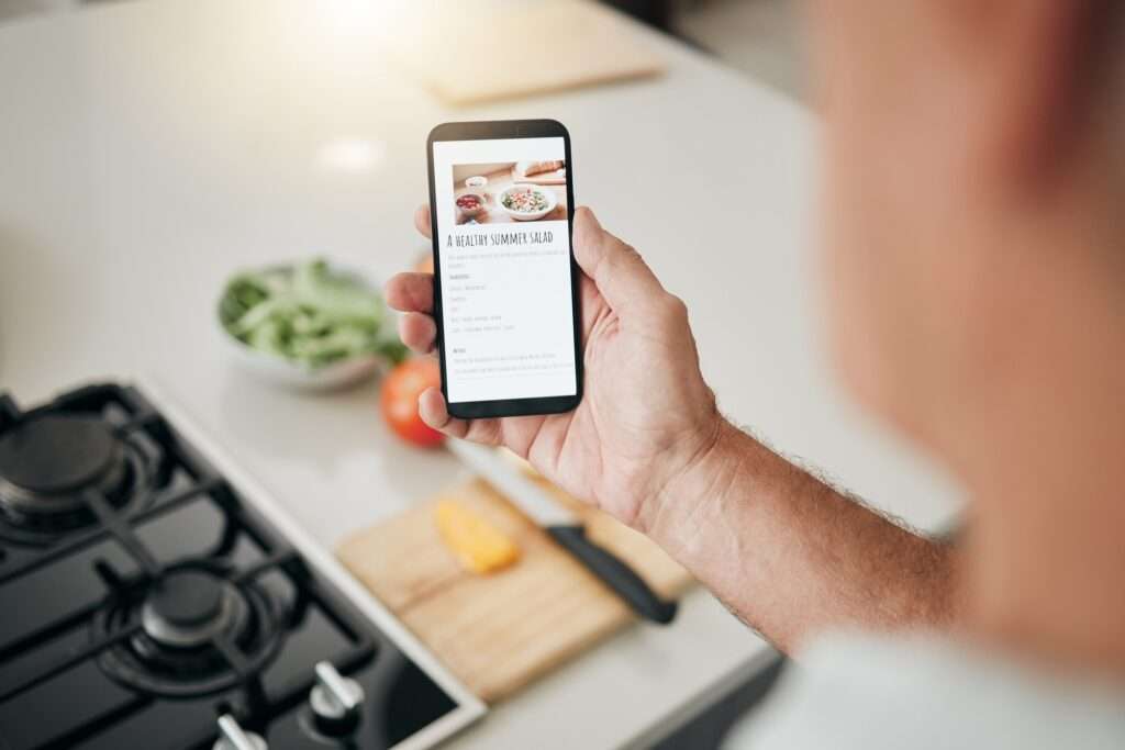 Webdesign Hannover - In einer Küche hält eine Person ein Smartphone, auf dem ein Rezept mit dem Titel „Ein gesunder Sommersalat“ angezeigt wird, mit Optionen in einem Dropdown-Menü. Im Hintergrund sind ein Herd, eine Schüssel mit Gemüse, eine Tomate und ein Schneidebrett mit geschnittenen Paprikaschoten und einem Messer zu sehen. Local SEO Agentur Hannover.