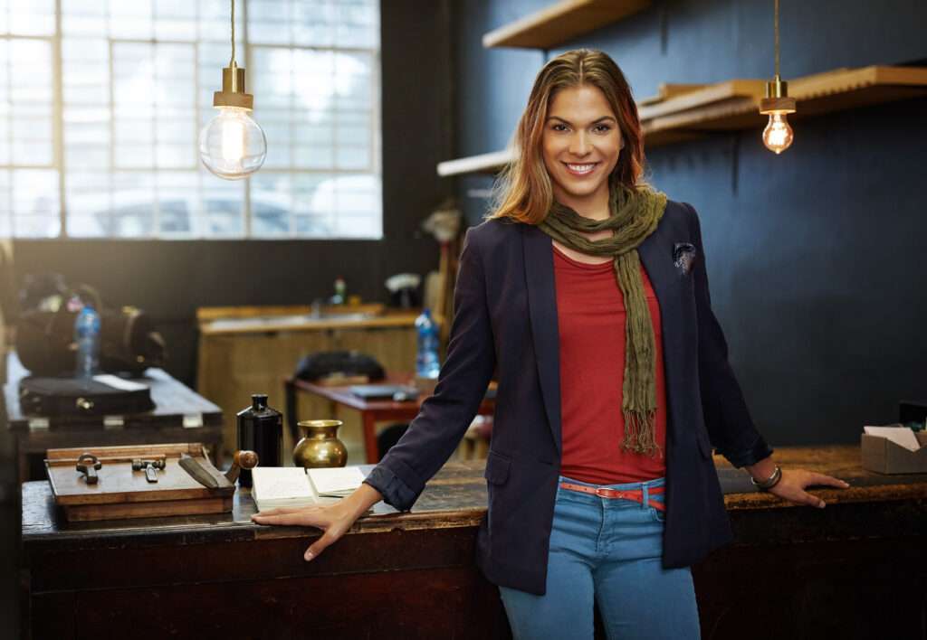 Webdesign Hannover - Eine Frau lehnt lächelnd an einer Holzwerkbank in einer Werkstatt und bereitet sich selbstbewusst darauf vor, ihr Unternehmen zu bestätigen. Sie trägt einen lässigen Blazer, einen Schal und Jeans; Regale, Werkzeuge und hängende Glühbirnen füllen den Raum des Kreativstudios. Local SEO Agentur Hannover.
