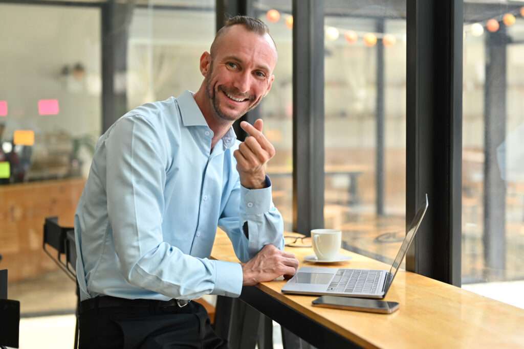 Webdesign Hannover - Ein Mann in einem hellblauen Hemd sitzt mit einem Laptop und einer Tasse Kaffee an einem Tresen am Fenster, lächelt und macht eine Handbewegung, während er in die Kamera blickt, als ob er bereit wäre, Ihnen bei der Bestätigung Ihres Unternehmens zu helfen. Local SEO Agentur Hannover.