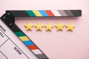 Webdesign Hannover - Eine Filmklappe mit farbigen Streifen liegt teilweise geöffnet auf einem rosa Untergrund. Davor sind fünf gelbe Sterne in einer Reihe angeordnet, die Top-Bewertungen suggerieren – perfekt, um zu visualisieren, wie sich Bewertungen auf Ihre Bewertungen auswirken können. Local SEO Agentur Hannover.