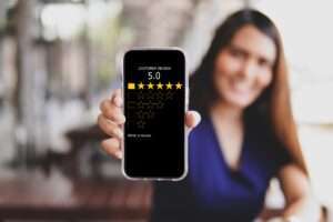Webdesign Hannover - Eine Person hält ein Smartphone, auf dem ein Kundenbewertungsbildschirm mit einer 5,0-Sterne-Bewertung angezeigt wird, wobei alle fünf Sterne hervorgehoben sind und ein unscharfer Hintergrund mit Sitzgelegenheiten im Freien zu sehen ist – perfekt für alle, die neugierig auf Bewertungen bei Google sind. Local SEO Agentur Hannover.