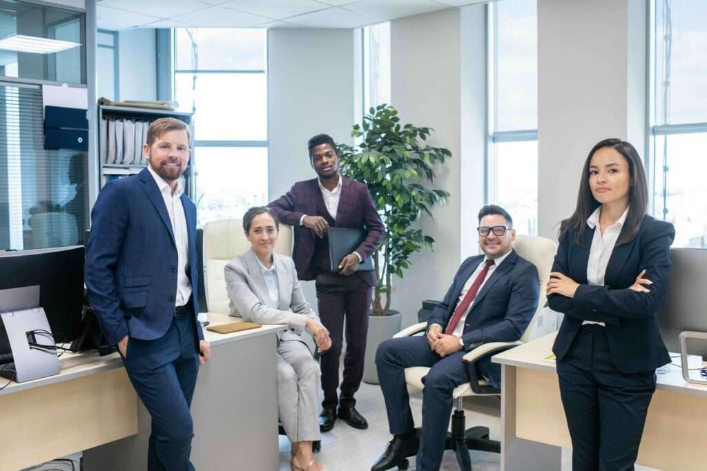 Webdesign Hannover - In einem eleganten Büro strahlen fünf Geschäftsleute in Business-Kleidung Selbstvertrauen und Ausgeglichenheit aus. Drei stehen, zwei sitzen. Eine große Pflanze und Schreibtische mit Computern bilden den Hintergrund. Dieses Bild fängt die Essenz des Unternehmenserfolgs ein, gesehen durch die Linse eines Wirtschaftsfotografen in Hannover. Local SEO Agentur Hannover.